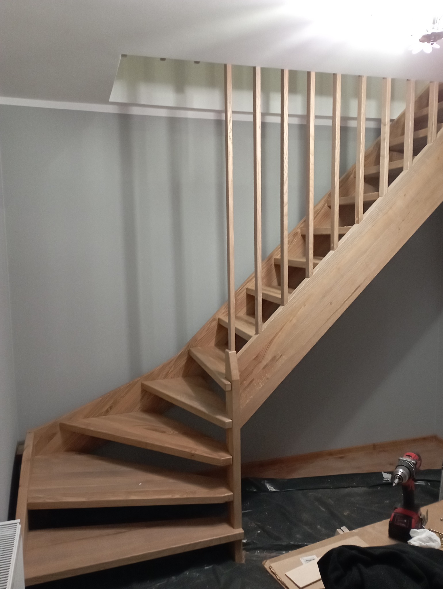 Jasne, drewniane schody spiralne z prostymi balustradami w trakcie montażu, widoczny wkrętak akumulatorowy i zabezpieczenie podłogi folią. Ujęcie z dołu.