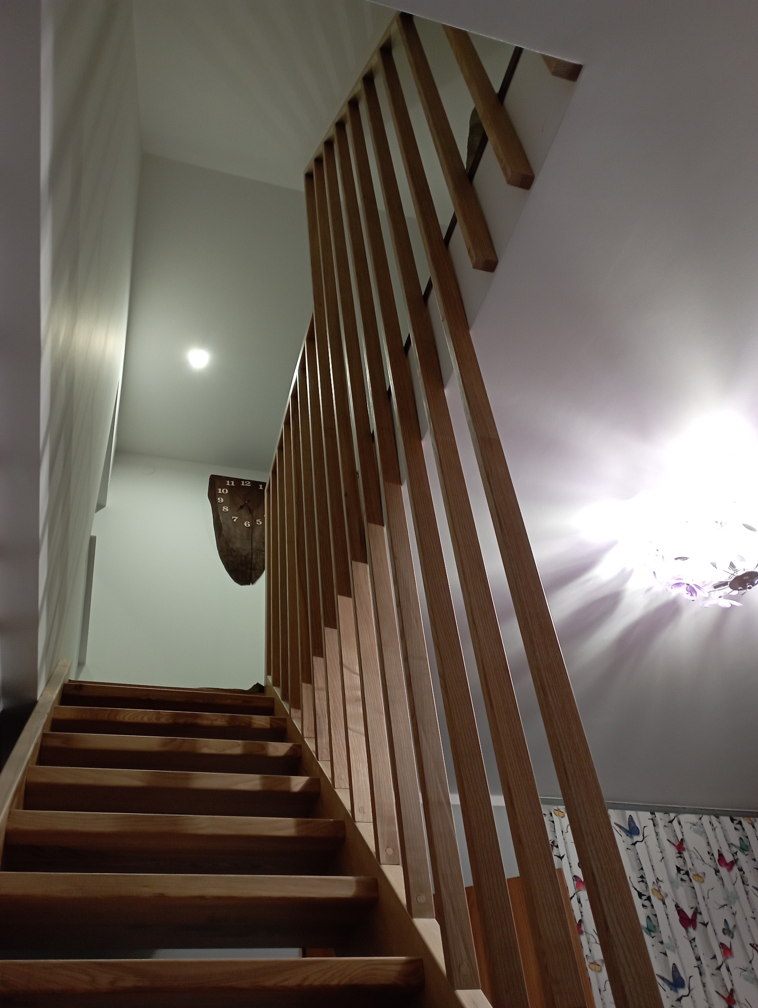 Drewniane schody z ażurową balustradą z pionowych listew, widok z dołu. Na ścianie drewniany zegar. W tle zasłony w motyle.