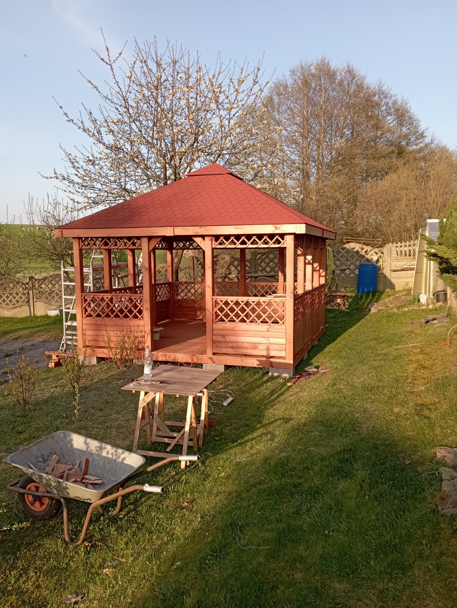 Drewniana altana z czerwonym dachem w ogrodzie, w trakcie montażu. Widoczny stół roboczy, taczka z drewnem i drabina. W tle drzewa i ogrodzenie.