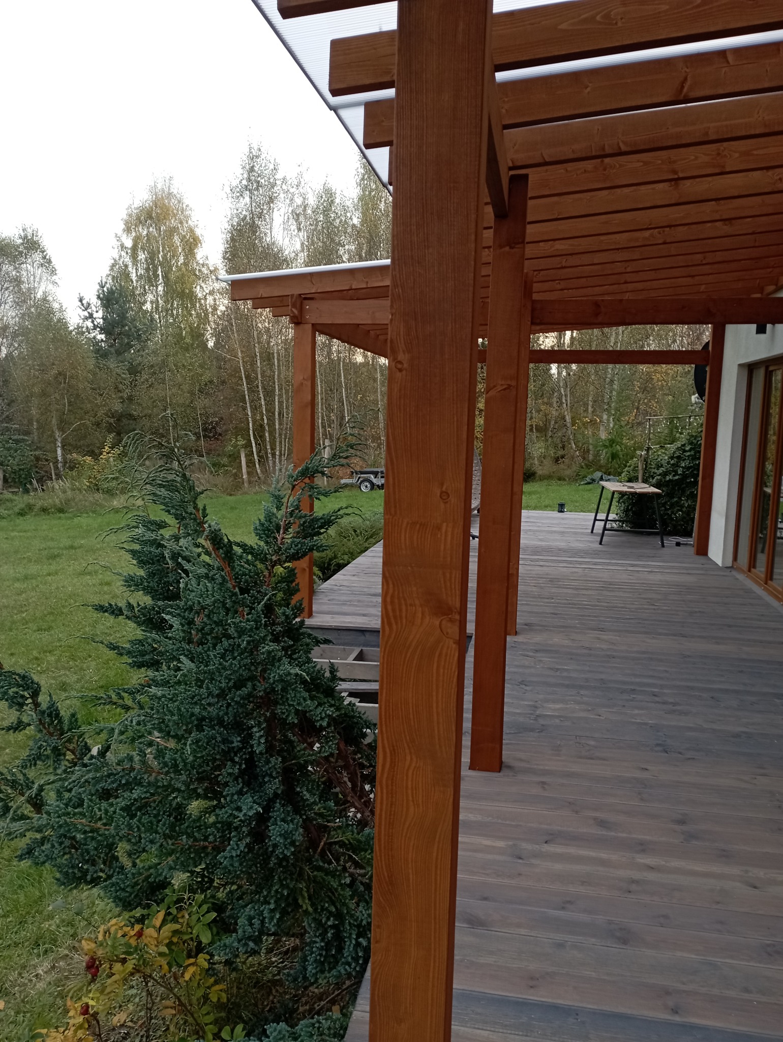 Drewniana pergola z zadaszeniem, widok z boku, z elementami zieleni w tle. Konstrukcja z belek w ciepłym odcieniu brązu, deski tarasowe w szarym kolorze.