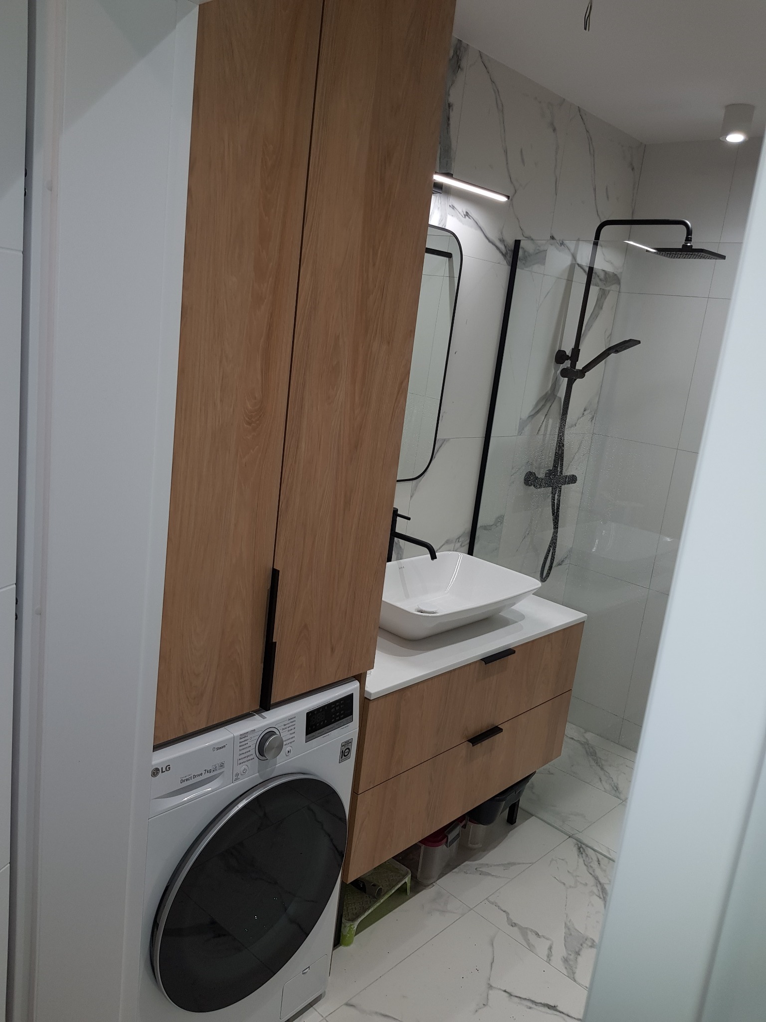 Nowoczesna łazienka z drewnianą szafką pod umywalką, białą umywalką nablatową, kabiną prysznicową z czarną armaturą i pralką LG w zabudowie.