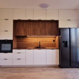 Nowoczesna kuchnia w Toruniu z białymi szafkami górnymi i dolnymi, drewnianym panelem nad blatem, czarnym piekarnikiem i lodówką side-by-side, oświetlona listwą LED pod szafkami.