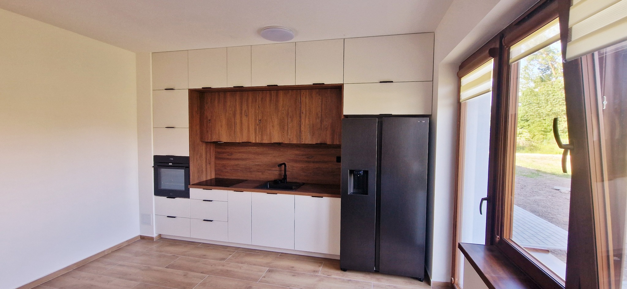 Nowoczesna kuchnia z białymi szafkami górnymi i dolnymi, drewnianym panelem nad blatem, czarnym zlewem i dużą, ciemną lodówką side-by-side, widok na zewnątrz przez okno.