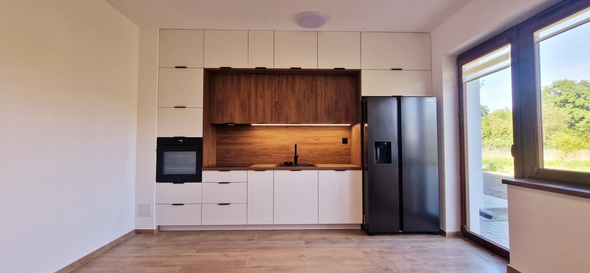 Nowoczesna kuchnia w Toruniu z białymi szafkami górnymi i dolnymi, drewnianym panelem nad blatem, czarnym piekarnikiem i lodówką side-by-side, oświetlona listwą LED pod szafkami.