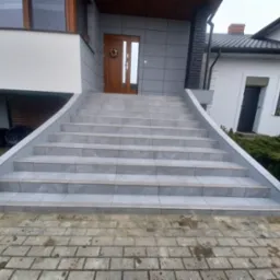 Zewnętrzne schody wejściowe do domu, wyłożone szarymi płytkami z antypoślizgowymi paskami, zakończone ozdobnymi, białymi kulami.