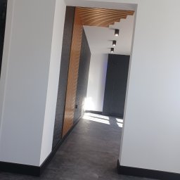 Kamil Lewandowski - Nowoczesne wnętrze z ciemnoszarą ścianą akcentową z drewnianymi panelami, listwami przypodłogowymi i podwieszanym sufitem z oświetleniem punktowym.