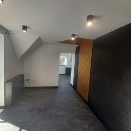 Kamil Lewandowski - Wnętrze po remoncie: czarna ściana z fakturą, drewniane lamele na suficie i ścianie, szara podłoga, nowoczesne oświetlenie punktowe.