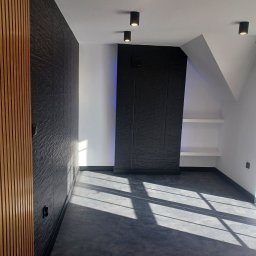Kamil Lewandowski - Nowoczesne wnętrze poddasza z czarną teksturowaną ścianą, drewnianymi lamelami po lewej stronie i rzędem punktowych lamp sufitowych, na podłodze widoczne promienie słoneczne.