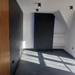 Nowoczesne wnętrze poddasza z czarną teksturowaną ścianą, drewnianymi lamelami po lewej stronie i rzędem punktowych lamp sufitowych, na podłodze widoczne promienie słoneczne.