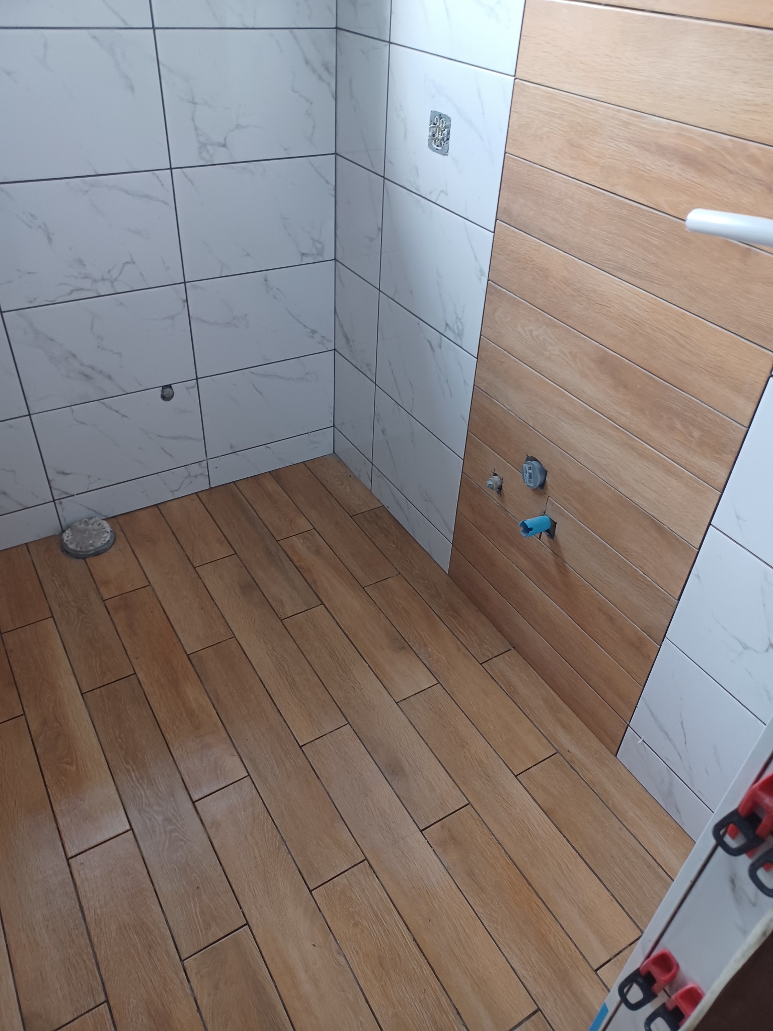 Wnętrze łazienki w trakcie prac wykończeniowych, widoczne płytki imitujące marmur na ścianach i drewnopodobne płytki na podłodze oraz fragment ściany z drewnopodobnymi płytkami, z wystającymi...