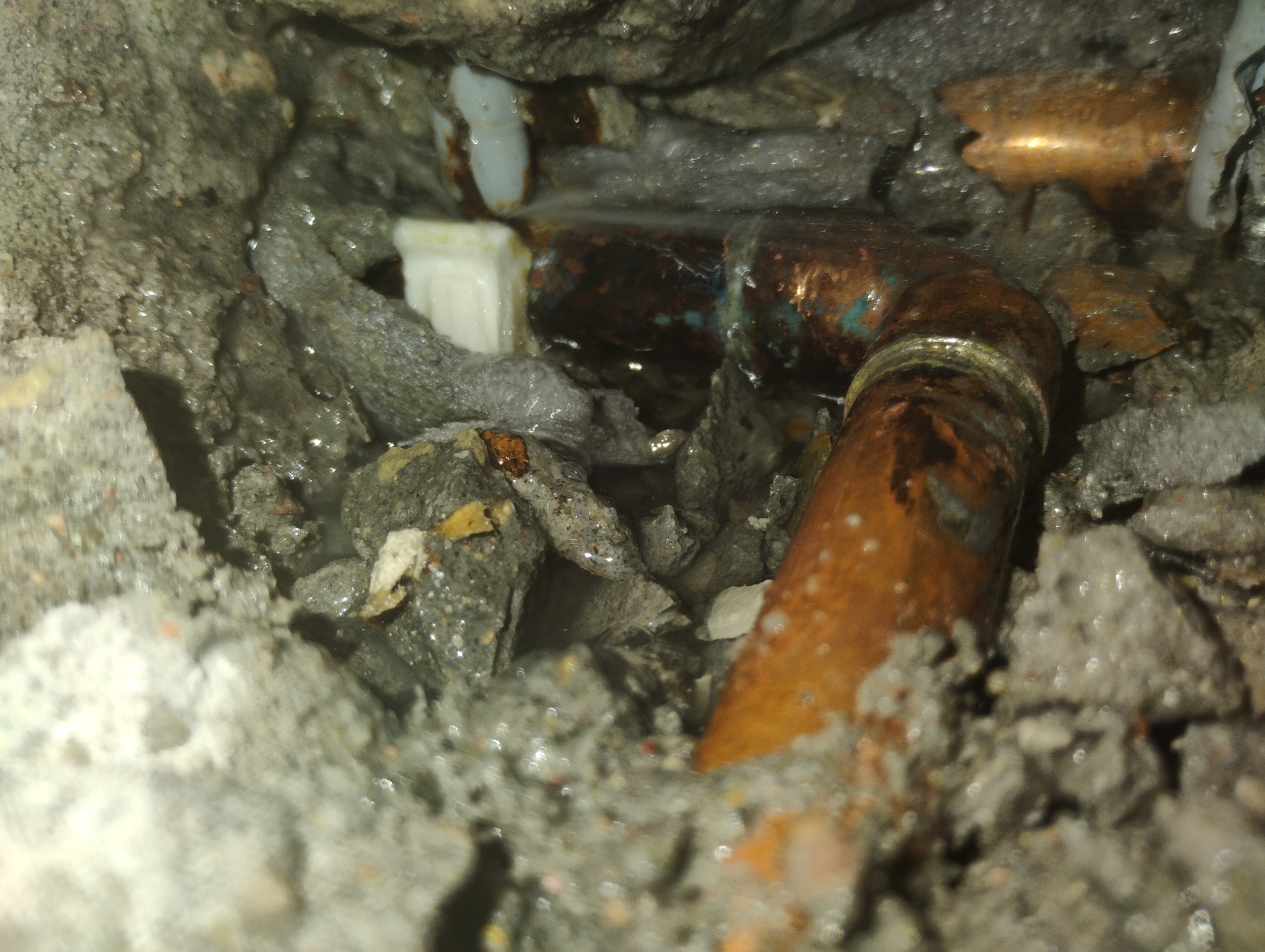 Fragment instalacji z miedzianą rurą, złączką PVC i betonowym gruzem, prawdopodobnie podczas naprawy lub wymiany, widoczne ślady wilgoci.