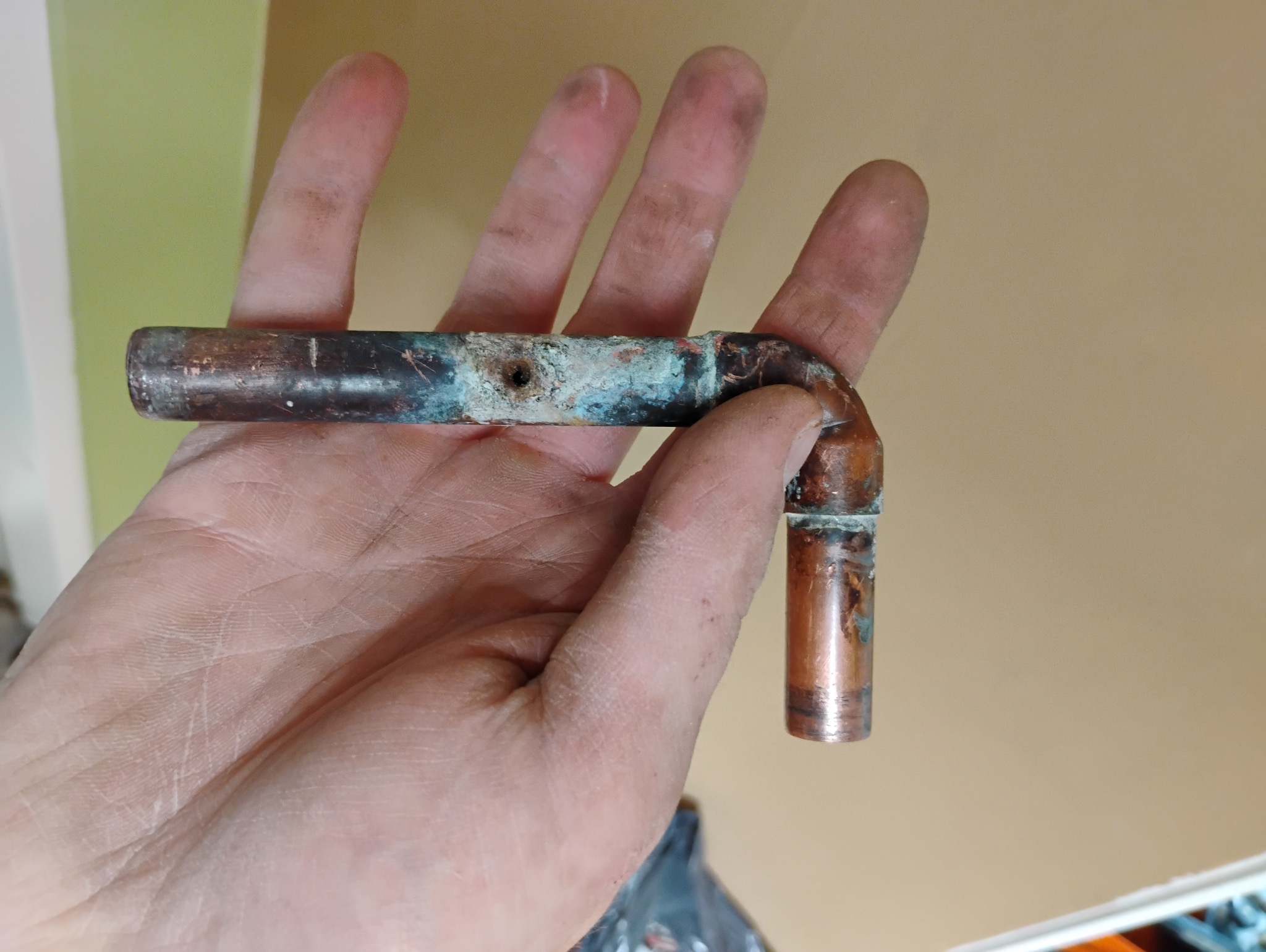 Miedziana rurka z otworem i śladami korozji trzymana w dłoni na tle ściany.