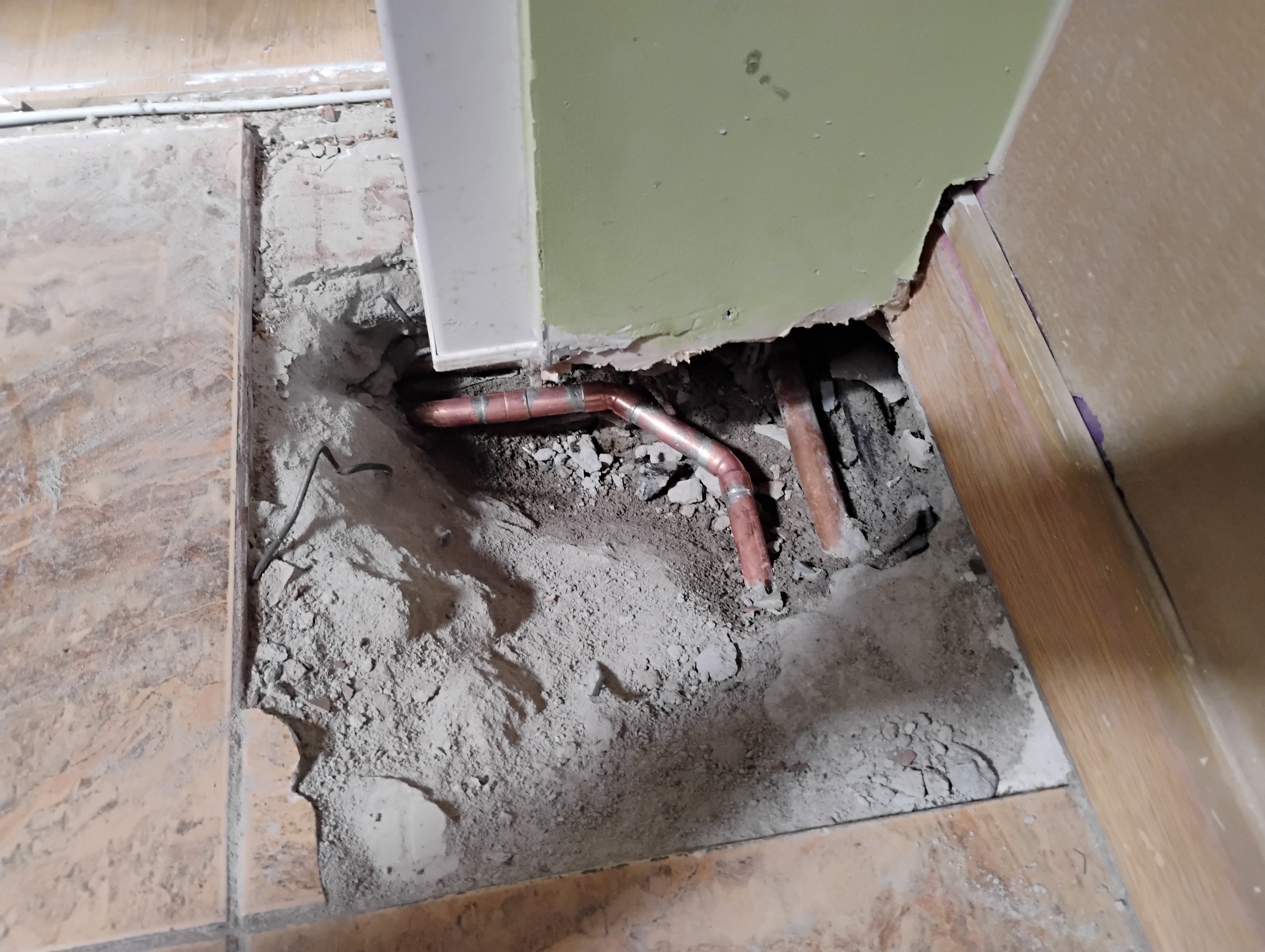 Odkryte miedziane rury instalacji wodnej w wykopanym otworze w podłodze z płytek, obok ściany z uszkodzonym tynkiem.