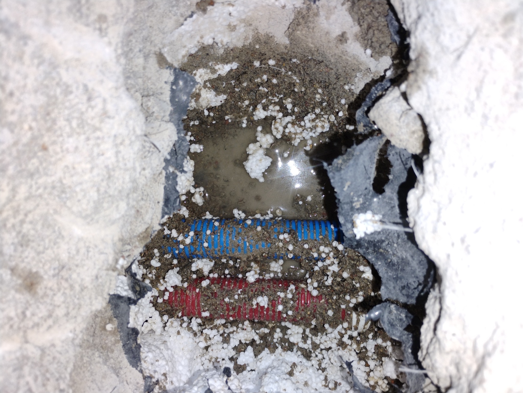 Odsłonięte, karbowane rury hydrauliczne (czerwona i niebieska) w wykuciu w ścianie, otoczone mokrym gruzem i białymi kulkami styropianu, częściowo zalane wodą.