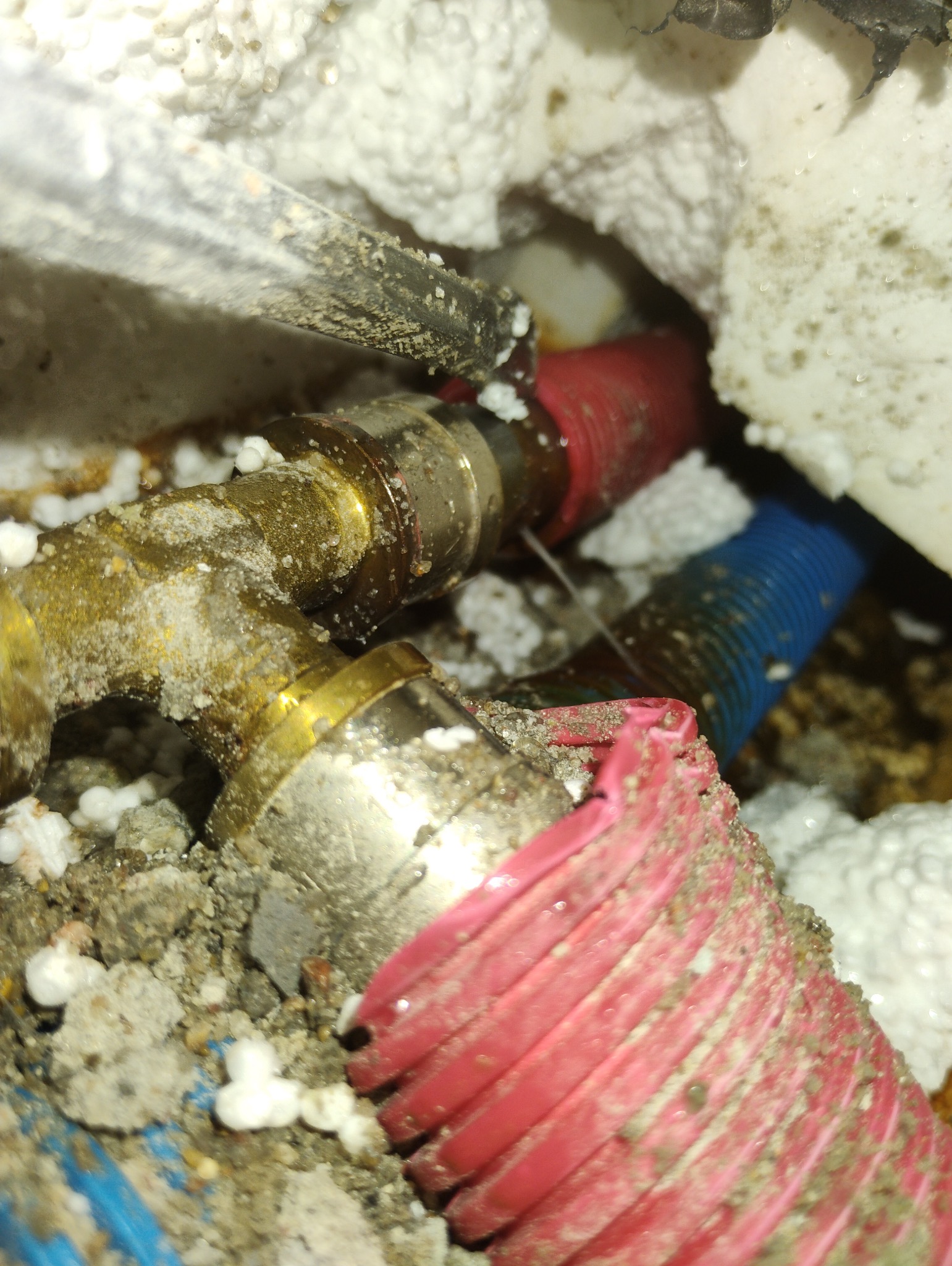 Wyciekająca woda z mosiężnego złącza instalacji hydraulicznej, otoczonego izolacją z białego styropianu i fragmentami rur w czerwonej i niebieskiej osłonie karbowanej, widoczne ślady korozji...