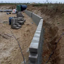 Budowa niskiego muru oporowego z betonowych bloczk&oacute;w na nier&oacute;wnym terenie, widoczne bloczki ułożone na palecie, czarna folia i zarys wykopu pod fundament.
