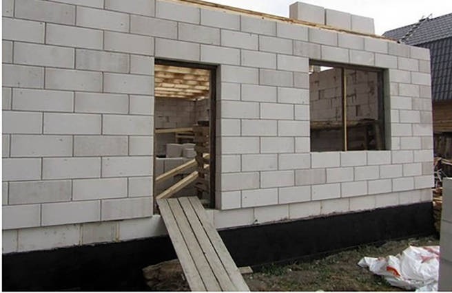 Wznoszenie ścian parterowego budynku z białych bloczków, widoczne otwory okienne i drzwiowe, przygotowane deski na wejście, w tle zarys dachu sąsiedniego budynku.