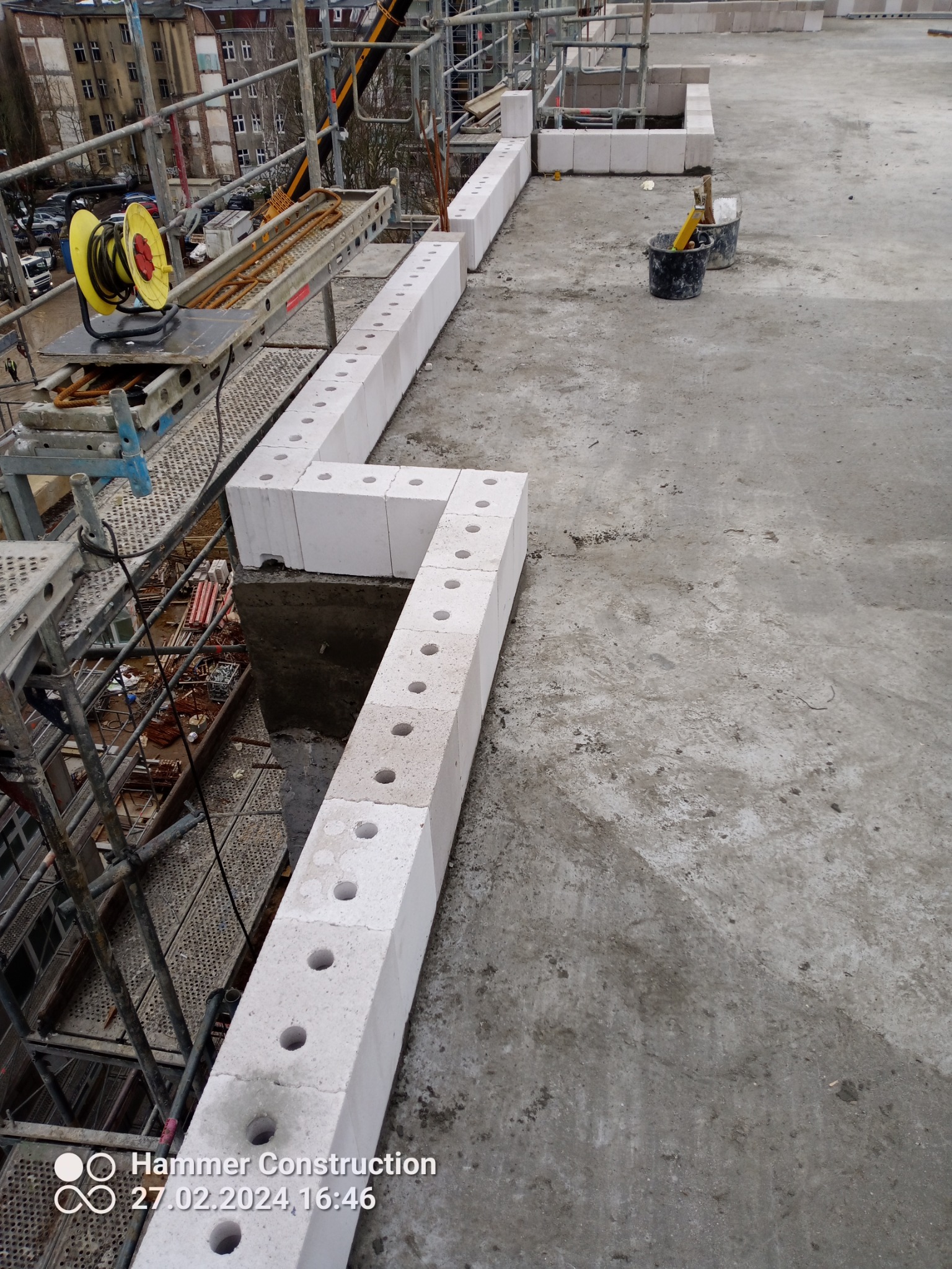 Wznoszone ściany z betonu komórkowego na budowie, widoczne rusztowania, zwijarka kabli i narzędzia w wiadrach na szarej betonowej powierzchni.