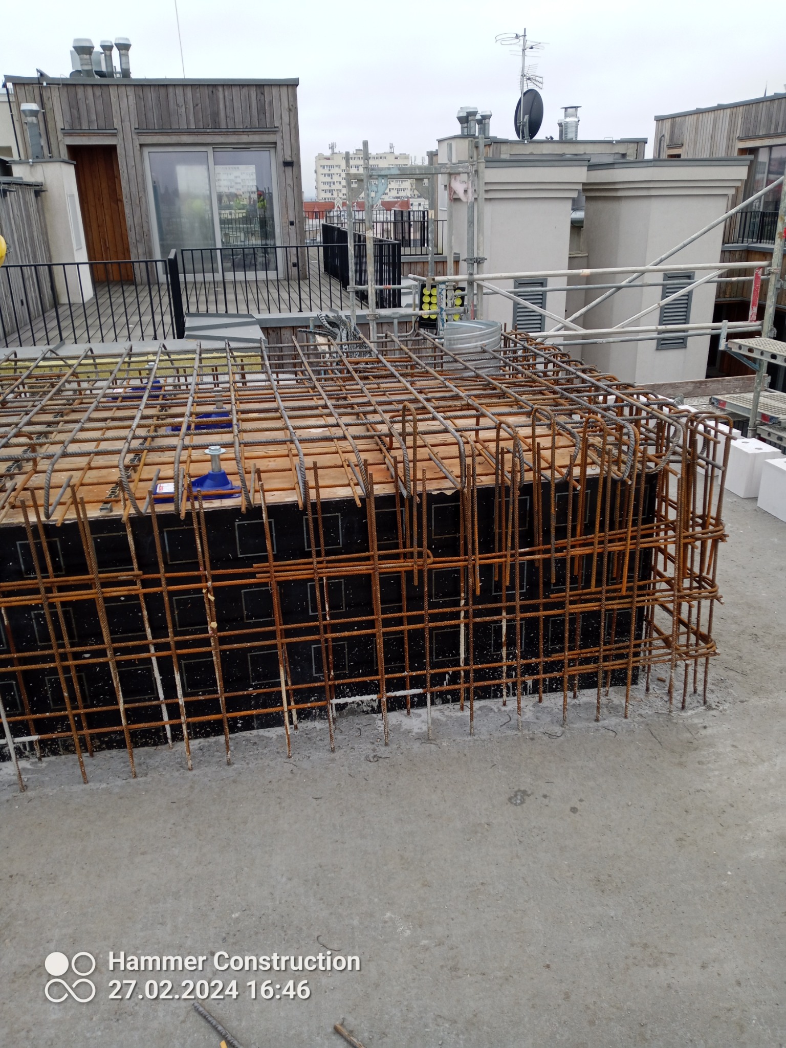 Zbrojenie szalunku fundamentu na dachu budynku wielorodzinnego, widoczne elementy konstrukcji i rusztowania w tle.
