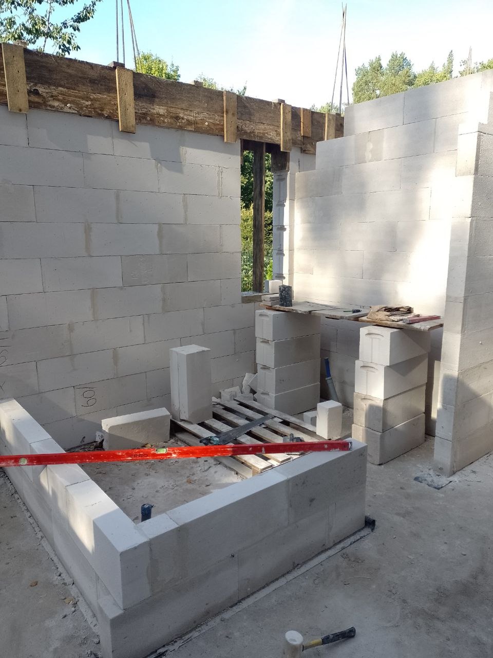 Wznoszenie ścian parteru z bloczków betonu komórkowego, widoczny poziomica, paleta, młotek i drewniane belki stropowe nad otworem okiennym.
