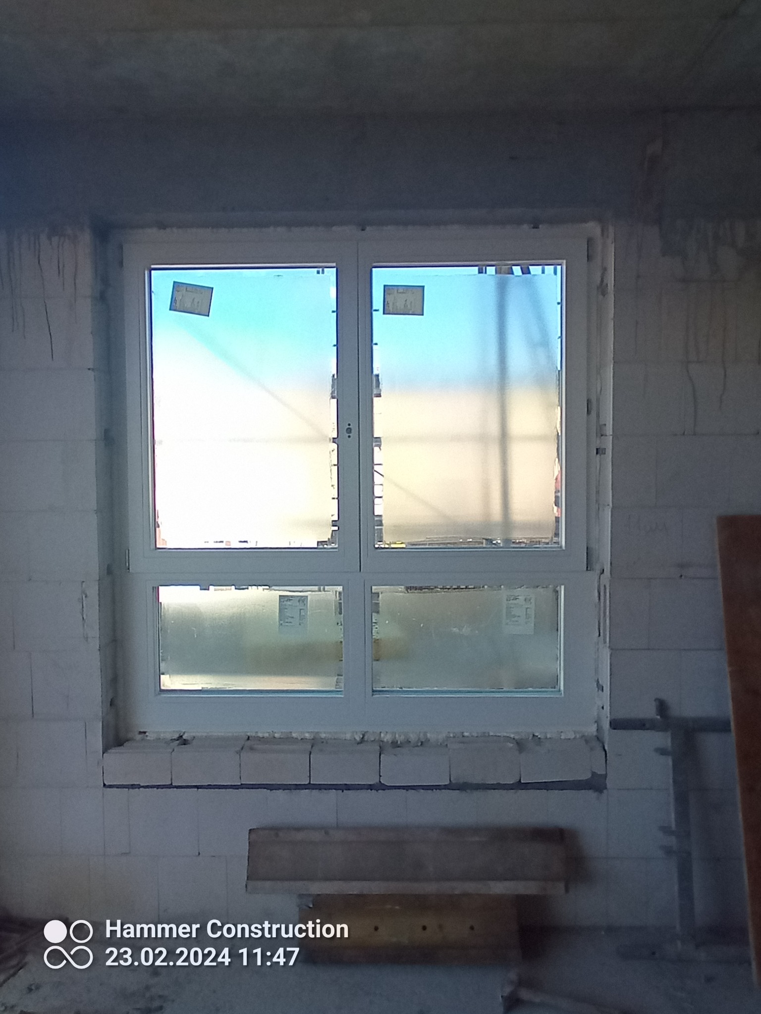 Nowe, białe okno PCV zamontowane w surowym murze z betonu komórkowego w trakcie budowy domu. Na szybach widoczne folie ochronne i naklejki producenta.