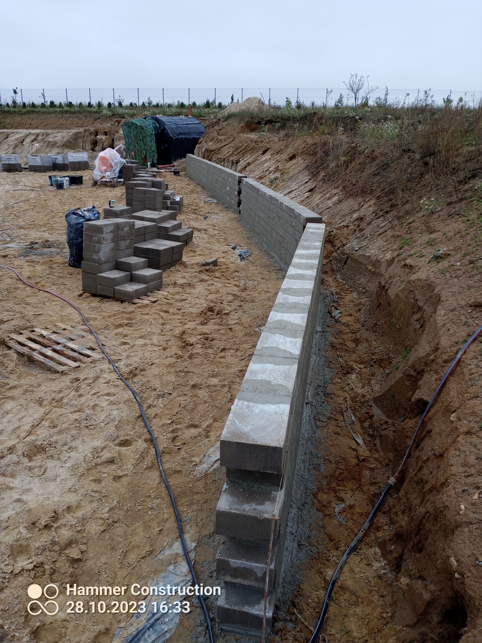Budowa niskiego muru oporowego z betonowych bloczków na nierównym terenie, widoczne bloczki ułożone na palecie, czarna folia i zarys wykopu pod fundament.