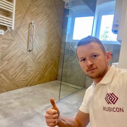 Rubicon - Ukończona łazienka z kabiną prysznicową typu walk-in, wykończona płytkami imitującymi drewno na ścianach i szarymi płytkami podłogowymi, z widocznym pracownikiem firmy Rubicon dającym kciuk w górę.