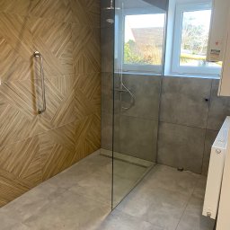 Rubicon - Nowoczesna kabina prysznicowa bez brodzika, z odpływem liniowym i szklaną ścianką, wykończona płytkami drewnopodobnymi w geometryczny wzór oraz szarymi płytkami wielkoformatowymi, z zamontowanym...