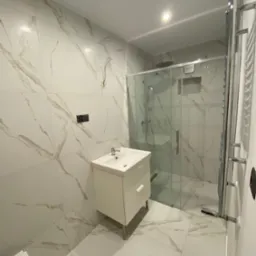 Nowoczesna łazienka z białymi płytkami imitującymi marmur, kabiną prysznicową ze szklanymi drzwiami i białą szafką pod umywalką; widoczne niedokończone podłączenie elektryczne