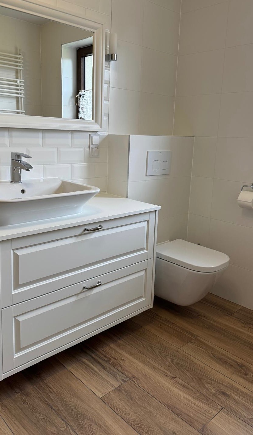 Jasna łazienka z drewnopodobną podłogą, białą umywalką nablatową z szafką, wiszącą toaletą i białymi płytkami typu metro na ścianach.