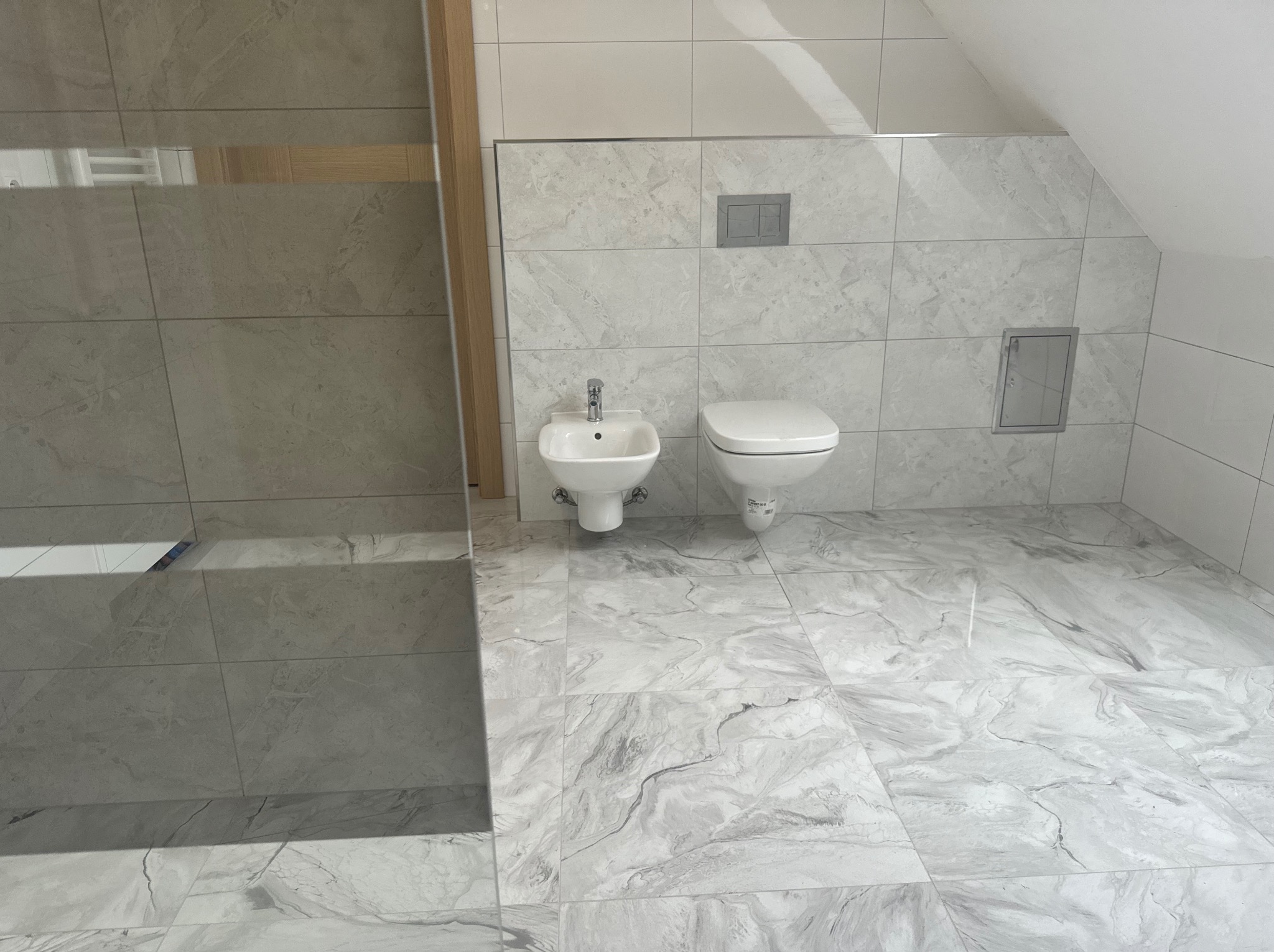 Wykończona łazienka na poddaszu z marmurowymi płytkami, wiszącą toaletą i bidetem, widoczny fragment kabiny prysznicowej z drewnianym obramowaniem.