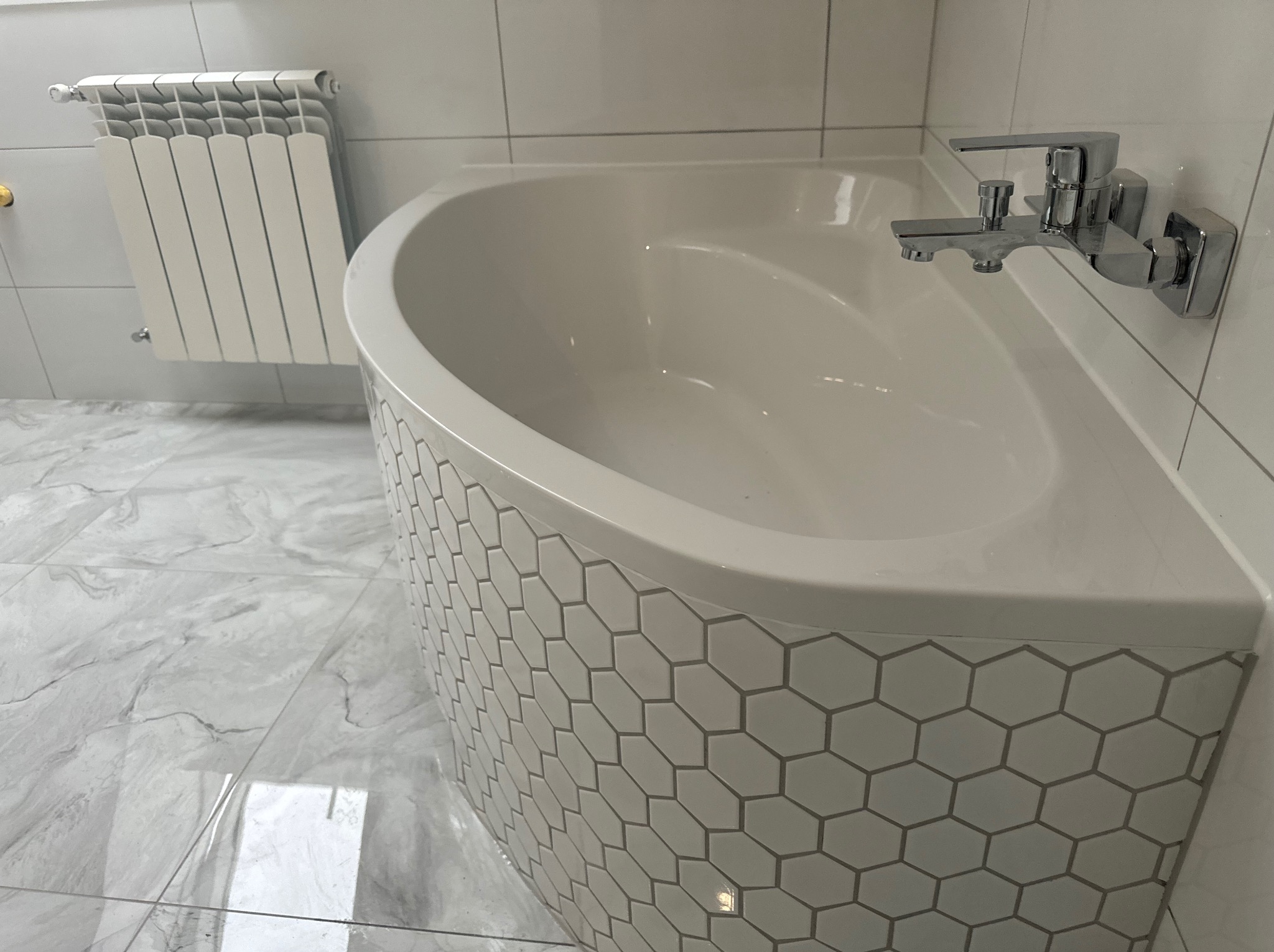 Biała wanna obudowana heksagonalnymi płytkami, chromowana bateria, marmurowe płytki podłogowe i biały grzejnik drabinkowy w łazience.