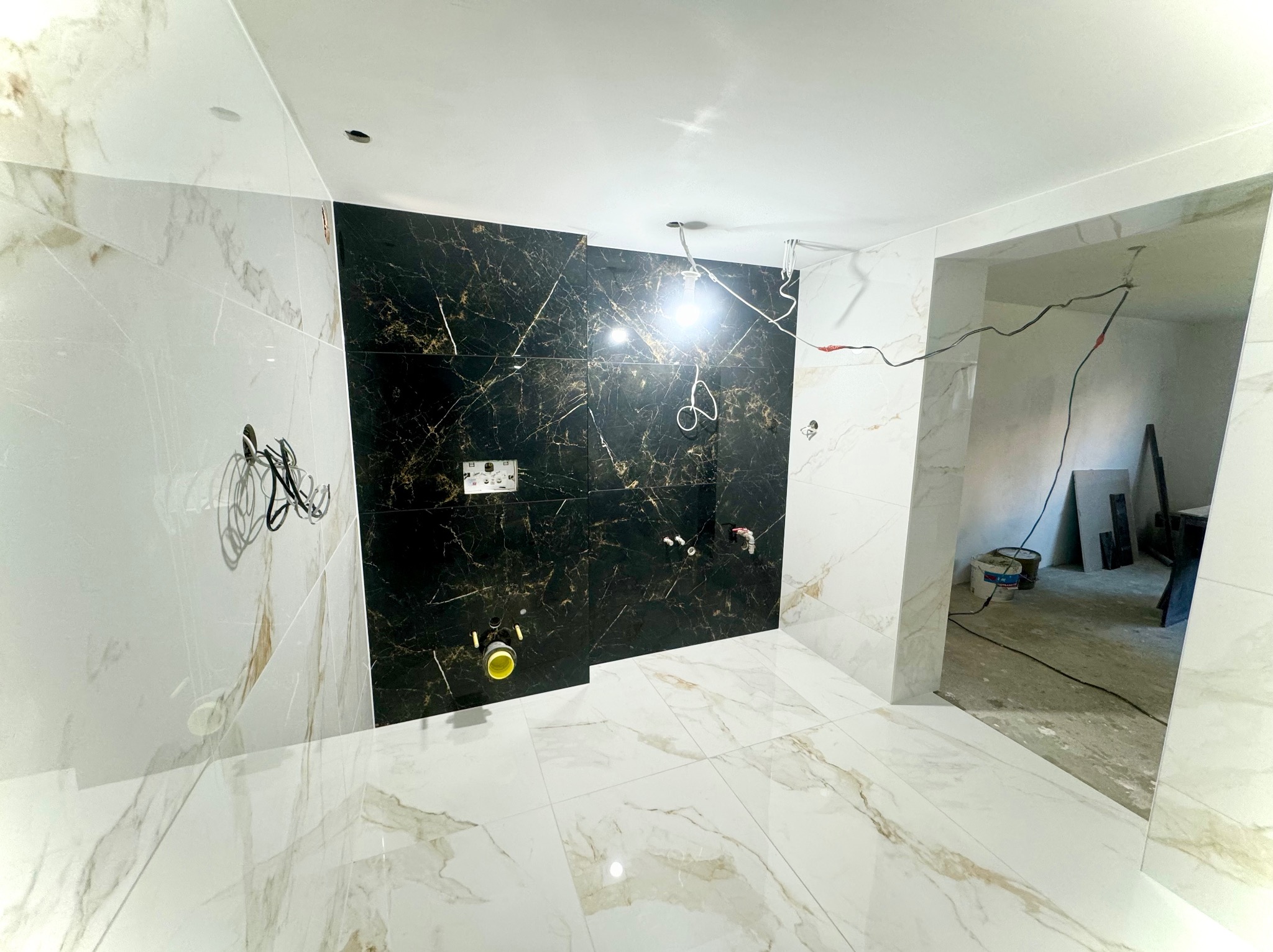 Wnętrze łazienki w trakcie prac wykończeniowych, widoczne czarne i białe płytki imitujące marmur na ścianach i podłodze, wystające przewody elektryczne, przygotowane podejścia wodno-kanalizacyjne.