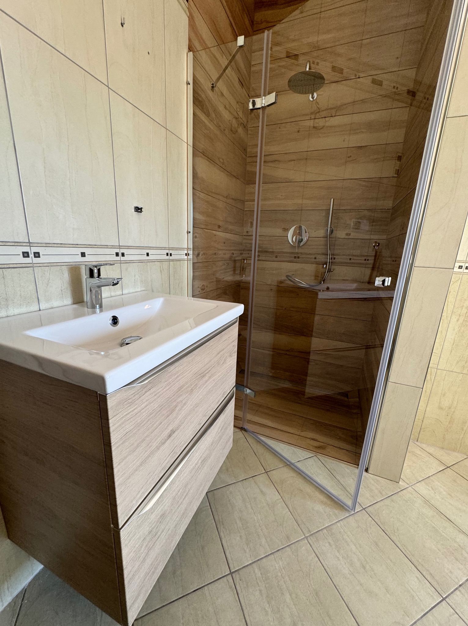 Nowoczesna łazienka z drewnopodobnymi płytkami w kabinie prysznicowej typu walk-in i szafką pod umywalką z dwiema szufladami o fakturze jasnego drewna.