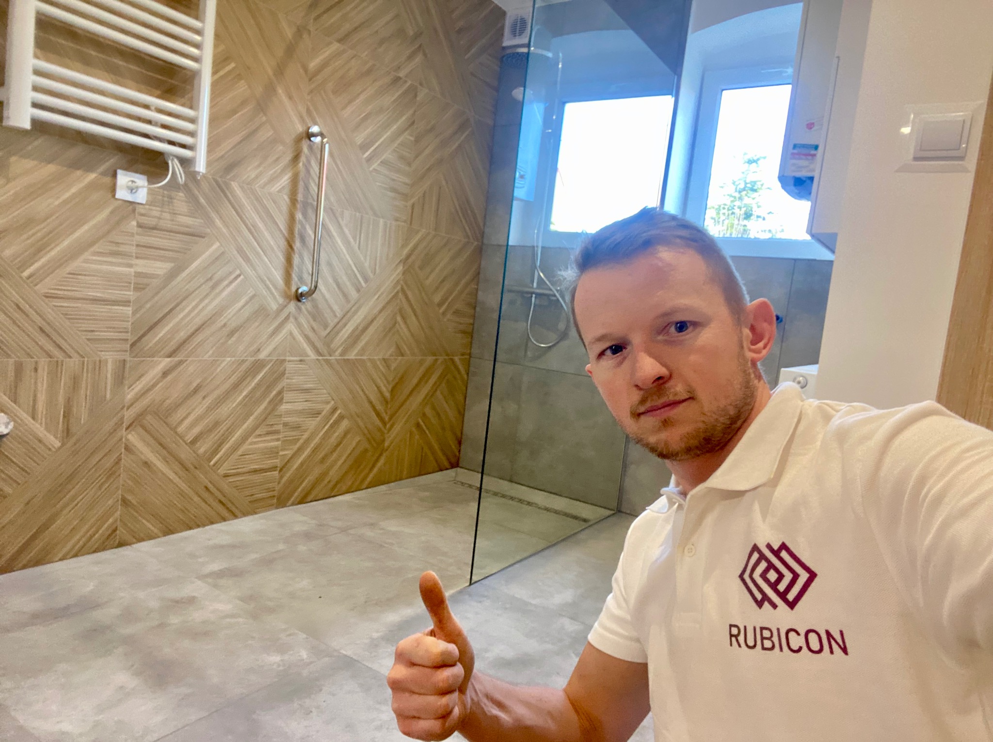 Ukończona łazienka z kabiną prysznicową typu walk-in, wykończona płytkami imitującymi drewno na ścianach i szarymi płytkami podłogowymi, z widocznym pracownikiem firmy Rubicon dającym kciuk w górę.