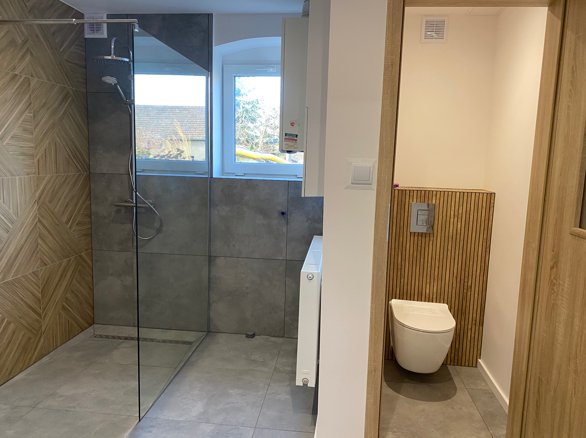 Nowoczesna łazienka z kabiną prysznicową typu walk-in z geometryczną mozaiką na ścianie, szarymi płytkami i toaletą podwieszaną z drewnianą boazerią w tle, widok zza framugi drzwi.