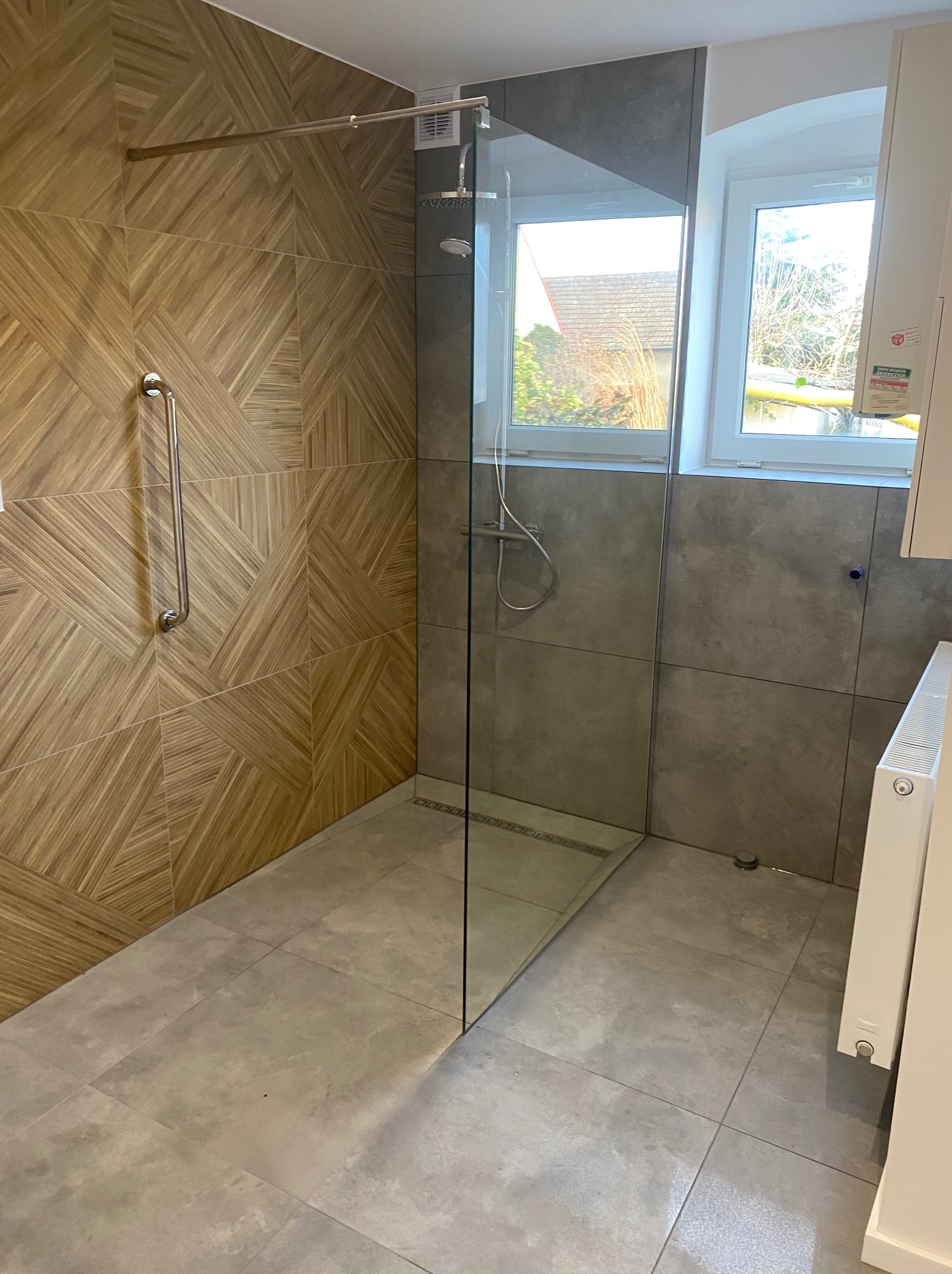 Nowoczesna kabina prysznicowa bez brodzika, z odpływem liniowym i szklaną ścianką, wykończona płytkami drewnopodobnymi w geometryczny wzór oraz szarymi płytkami wielkoformatowymi, z zamontowanym...