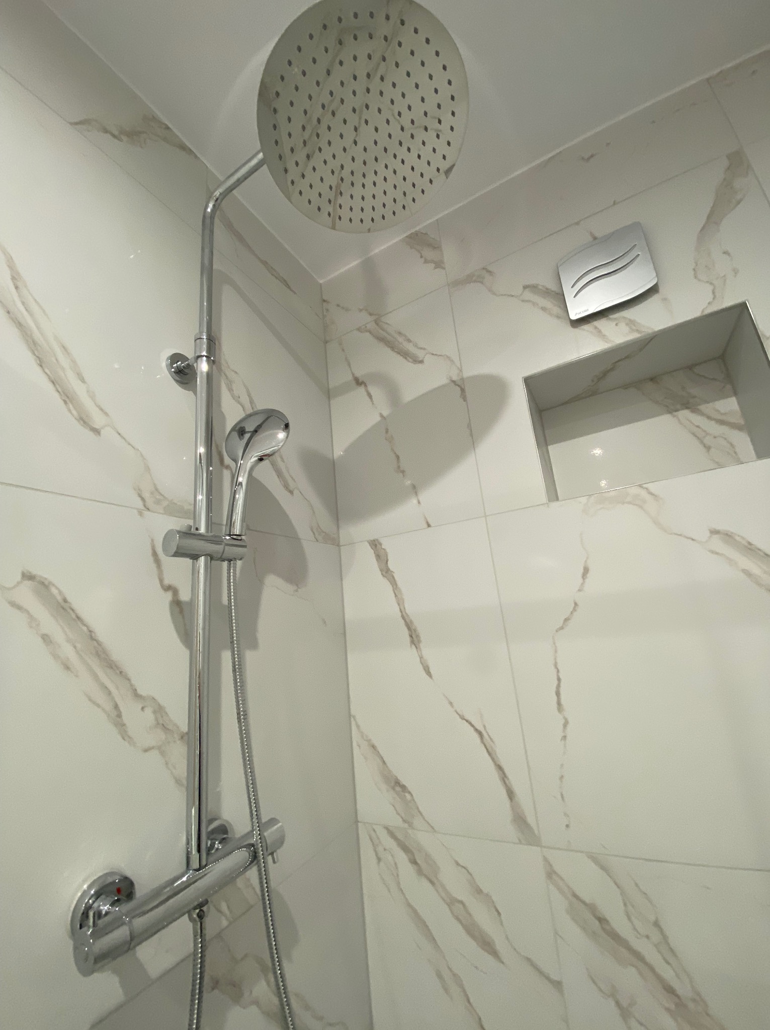 Nowoczesna kabina prysznicowa z chromowaną deszczownicą i słuchawką, białe płytki imitujące marmur, wnęka w ścianie i wentylator.