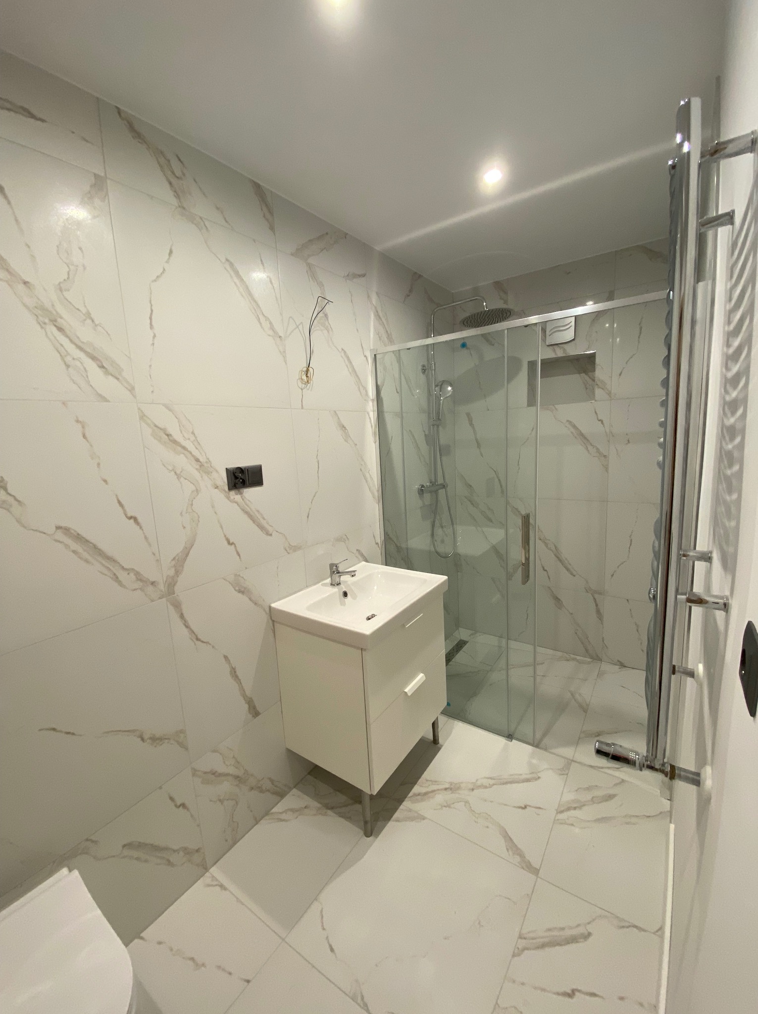 Nowoczesna łazienka z białymi płytkami imitującymi marmur, kabiną prysznicową ze szklanymi drzwiami i białą szafką pod umywalką; widoczne niedokończone podłączenie elektryczne