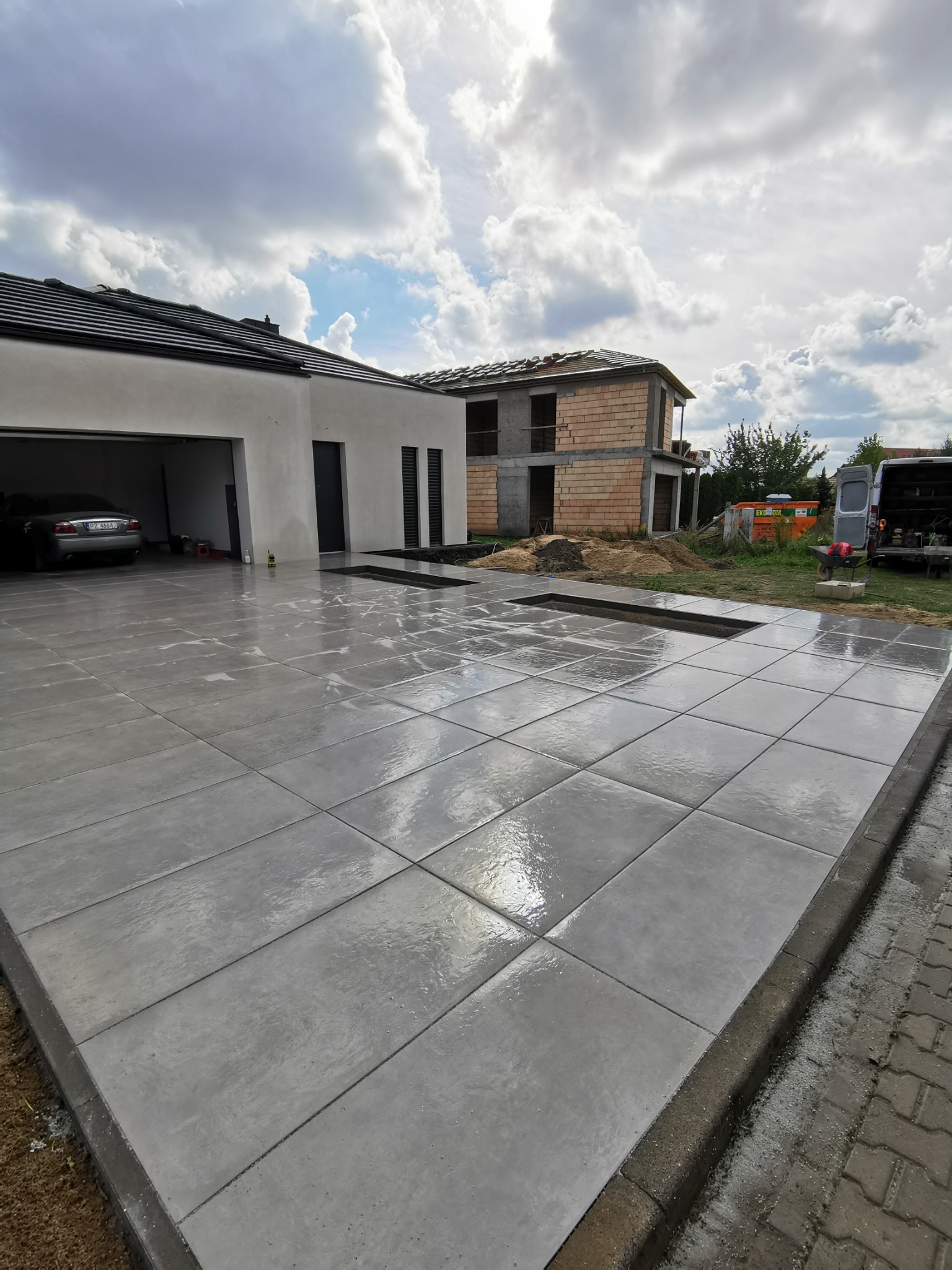 Świeżo ułożona, mokra, betonowa nawierzchnia z dużych płyt na podjeździe do garażu nowoczesnego domu, w tle budynek w stanie surowym otwartym i samochód osobowy.