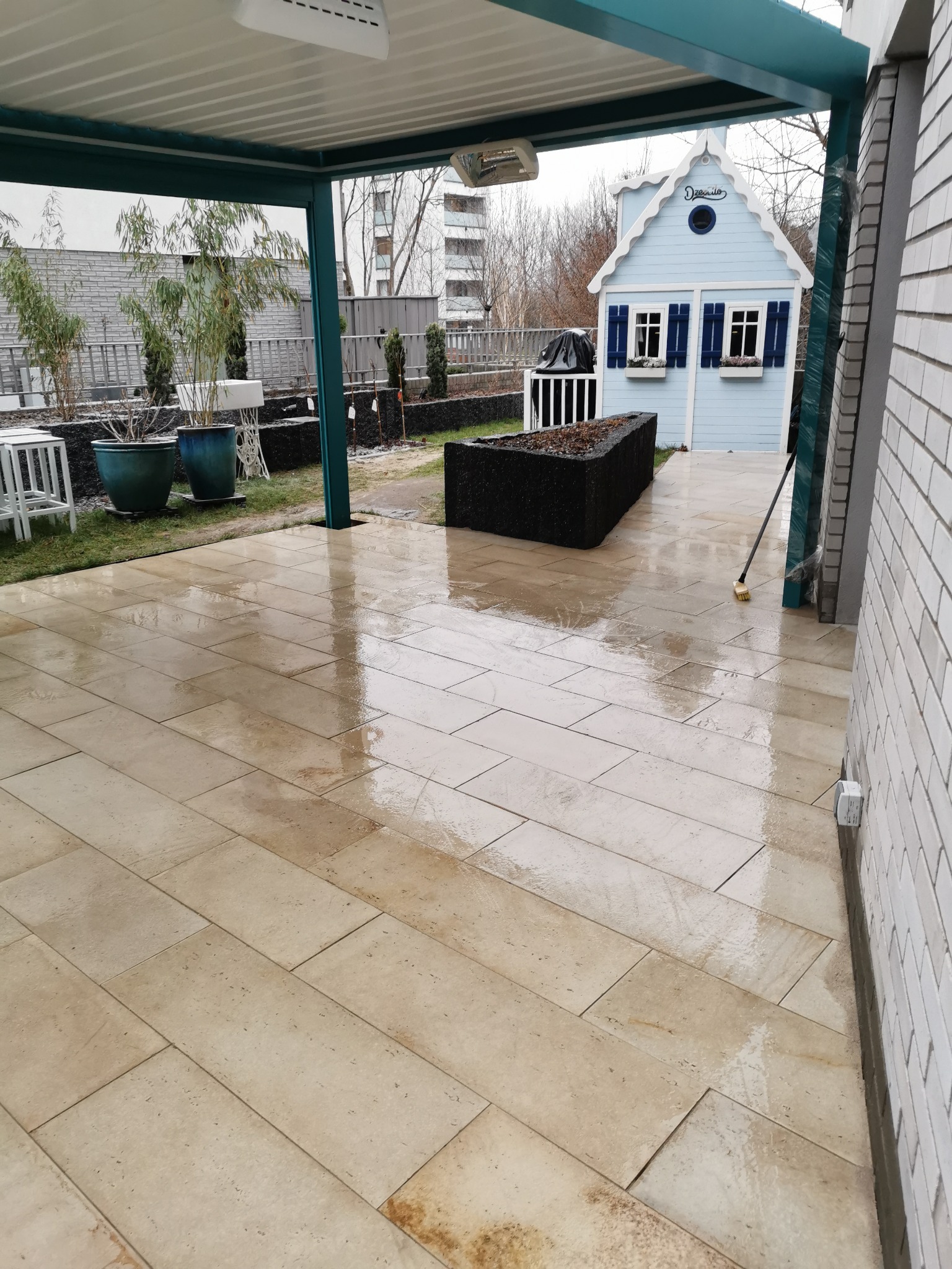 Świeżo ułożona, mokra, beżowa nawierzchnia z prostokątnych płyt, ułożonych w jodełkę, z widocznym fragmentem niebieskiego domku dla dzieci i roślinami w tle.