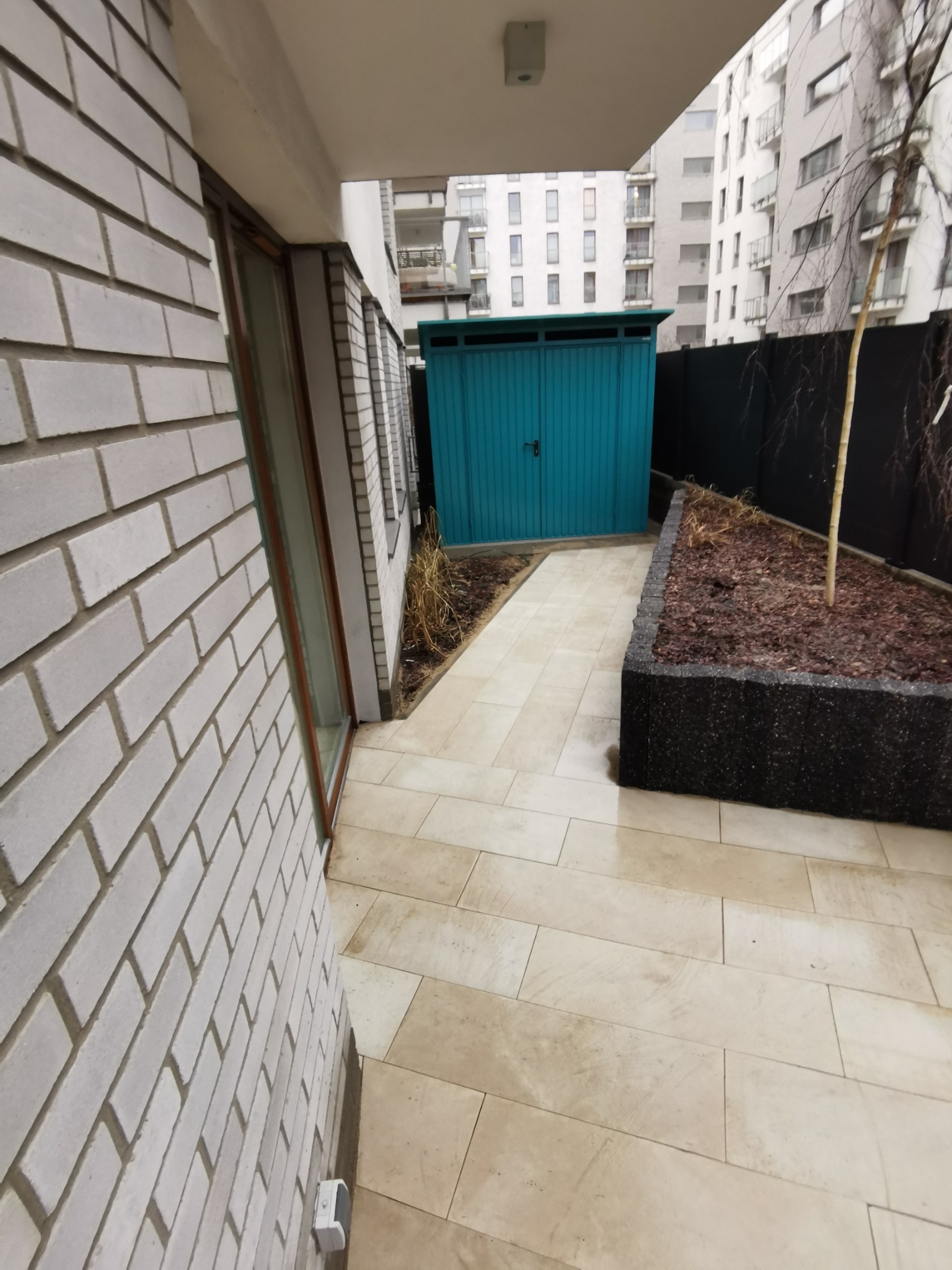 Ułożona jasna kostka brukowa na posesji z widocznym turkusowym schowkiem na narzędzia, donicą z korą i młodym drzewkiem.