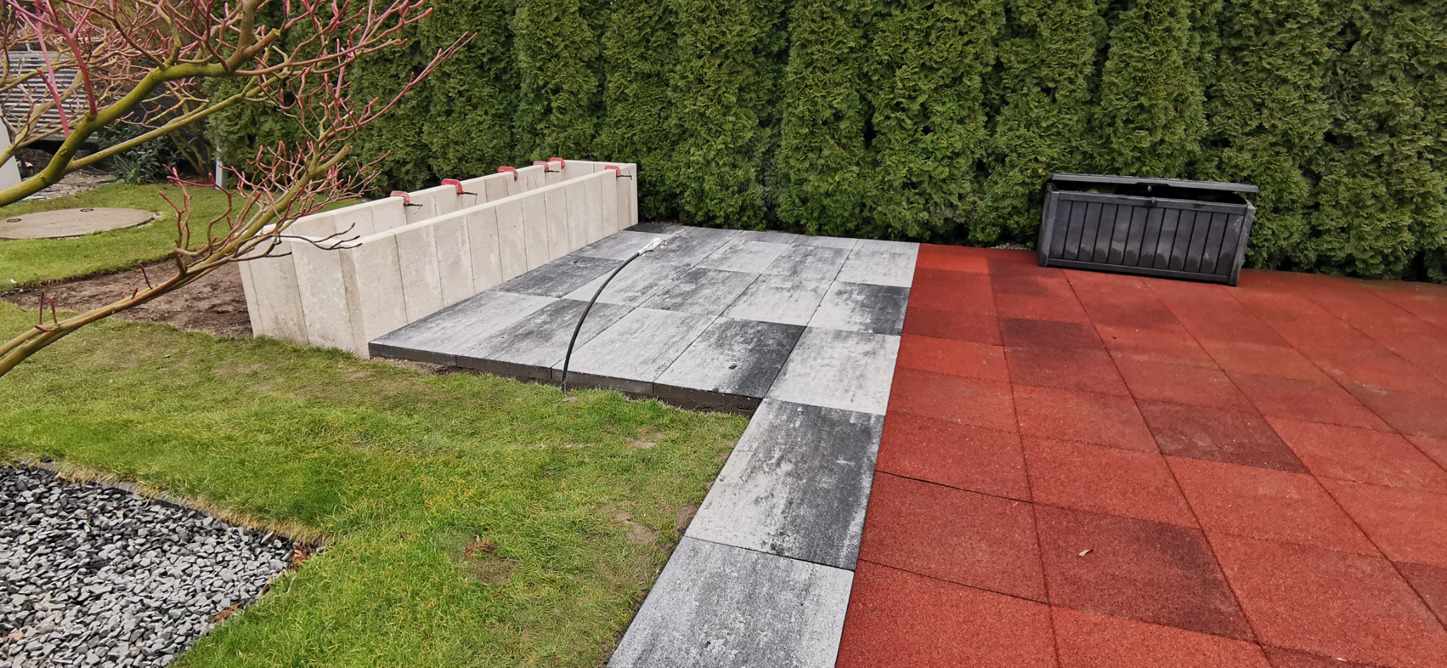 Fragment ogrodu z ułożoną szarą kostką brukową, przylegającą do czerwonej nawierzchni gumowej i betonowej konstrukcji, na tle zielonych tui.