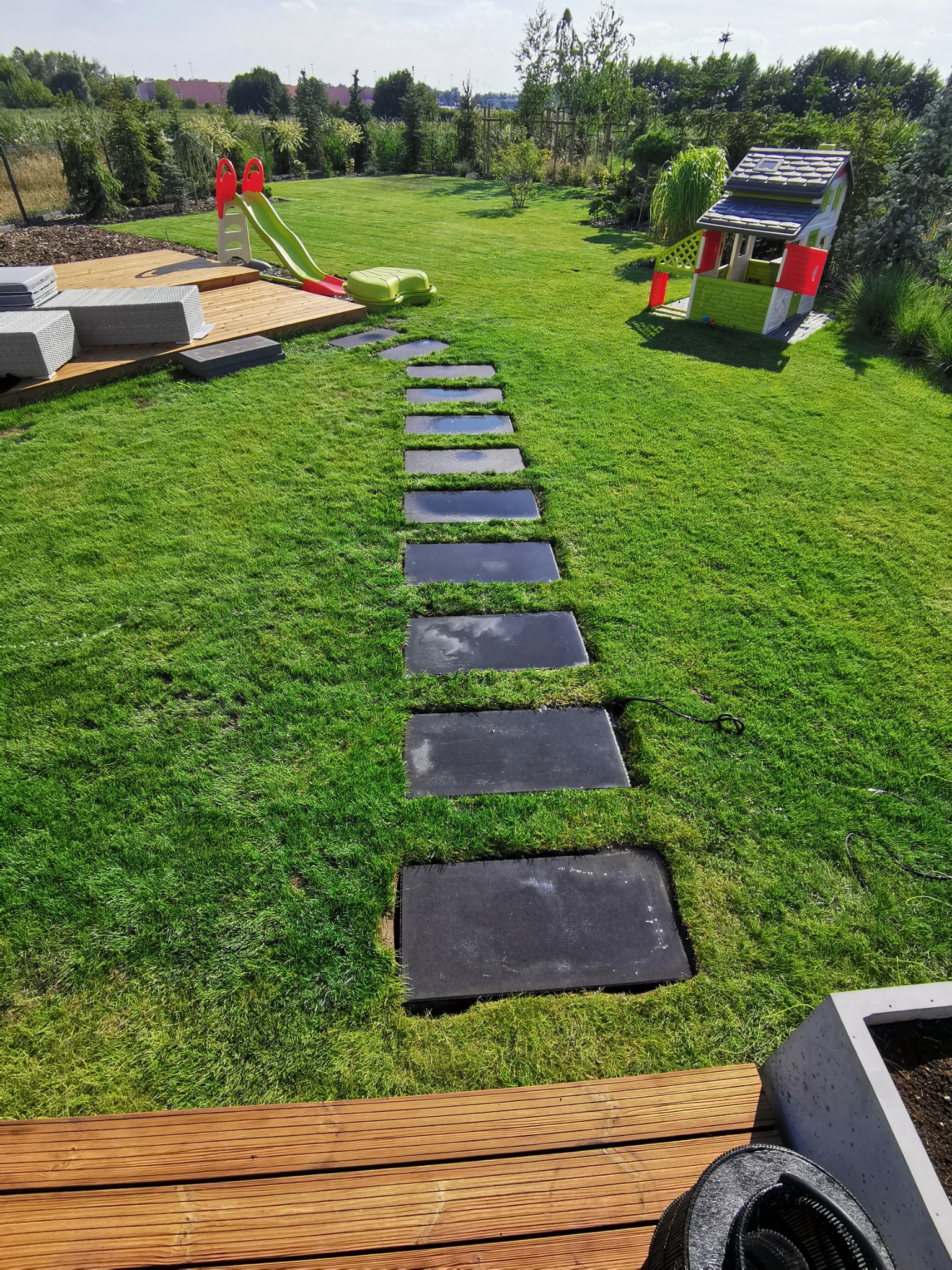 Ścieżka z ciemnych płyt ułożona w trawniku, prowadząca od drewnianego tarasu w stronę placu zabaw z domkiem i zjeżdżalnią, w tle zielony ogród.