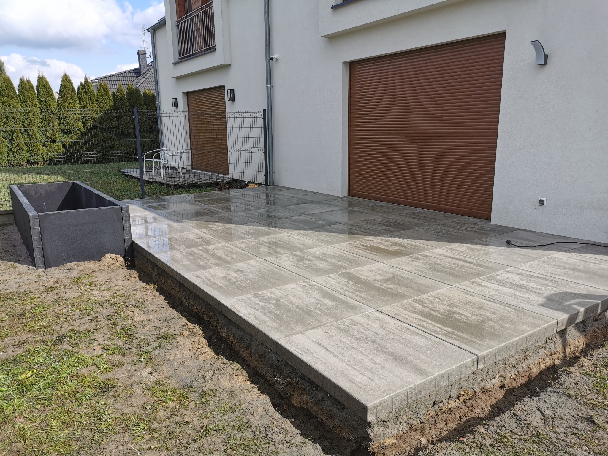 Świeżo ułożona, mokra, szara kostka brukowa na tarasie przed nowoczesnym domem z brązowymi bramami garażowymi, widoczne wykopane obrzeża i czarna donica na rośliny.