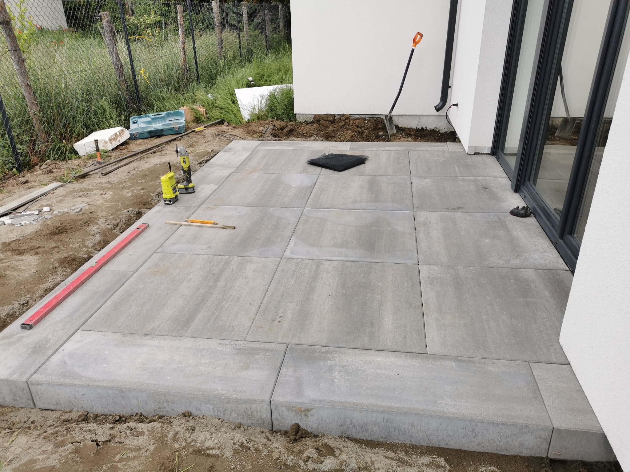 Niemal ukończony taras z szarej kostki brukowej w Poznaniu, widoczne narzędzia, poziomica i szpadel oparty o białą ścianę budynku.