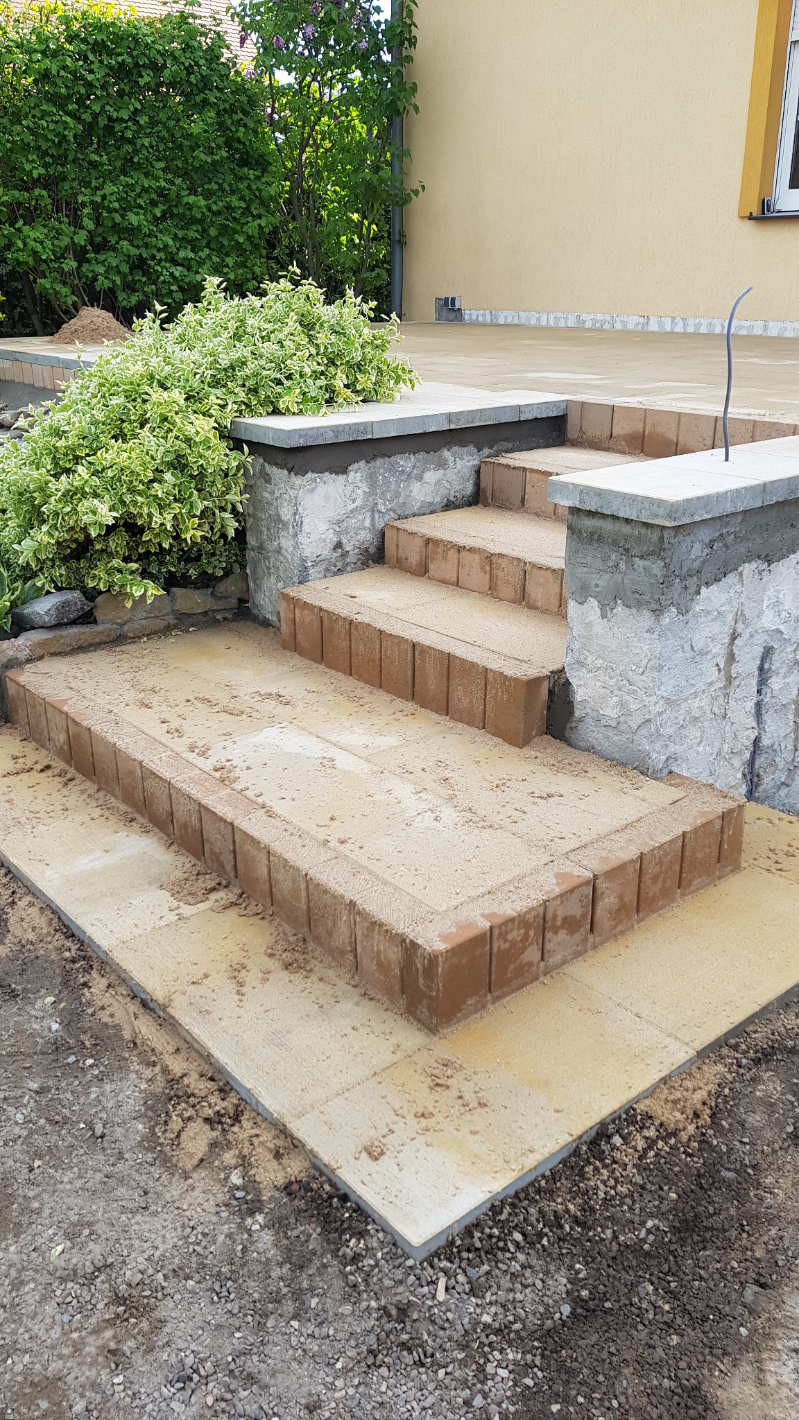 Schody z kostki brukowej w trakcie budowy, z widocznymi elementami konstrukcyjnymi i zaprawą, otoczone zielenią i częściowo wykończonym terenem.