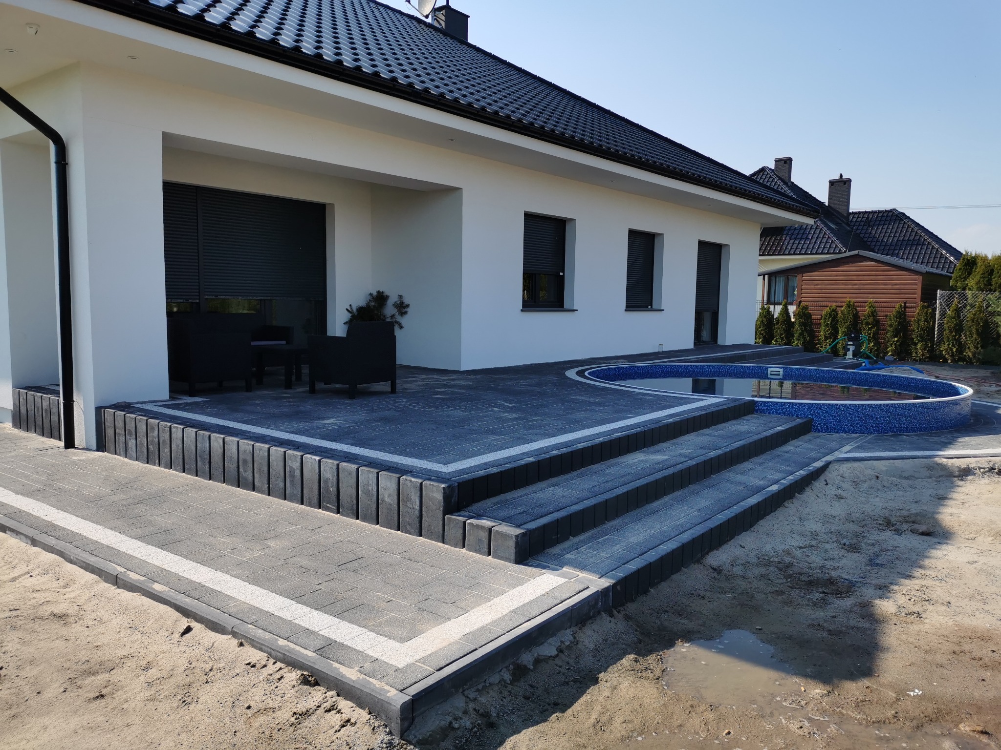 Nowoczesny taras z ciemnej kostki brukowej z jasnymi fugami, prowadzący do okrągłego basenu przy białym domu z czarną dachówką.