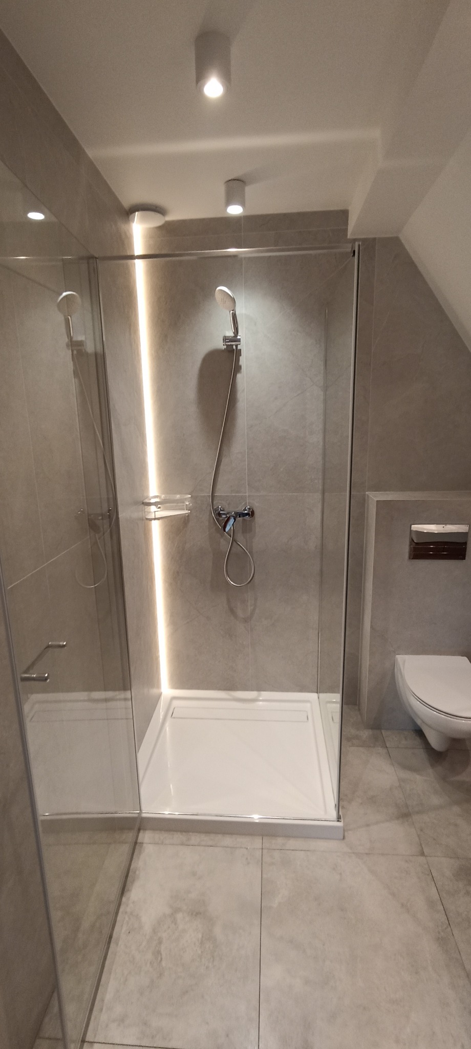 Nowoczesna kabina prysznicowa z oświetleniem LED w wyremontowanej łazience, szare płytki imitujące kamień, biały brodzik i toaleta w tle.