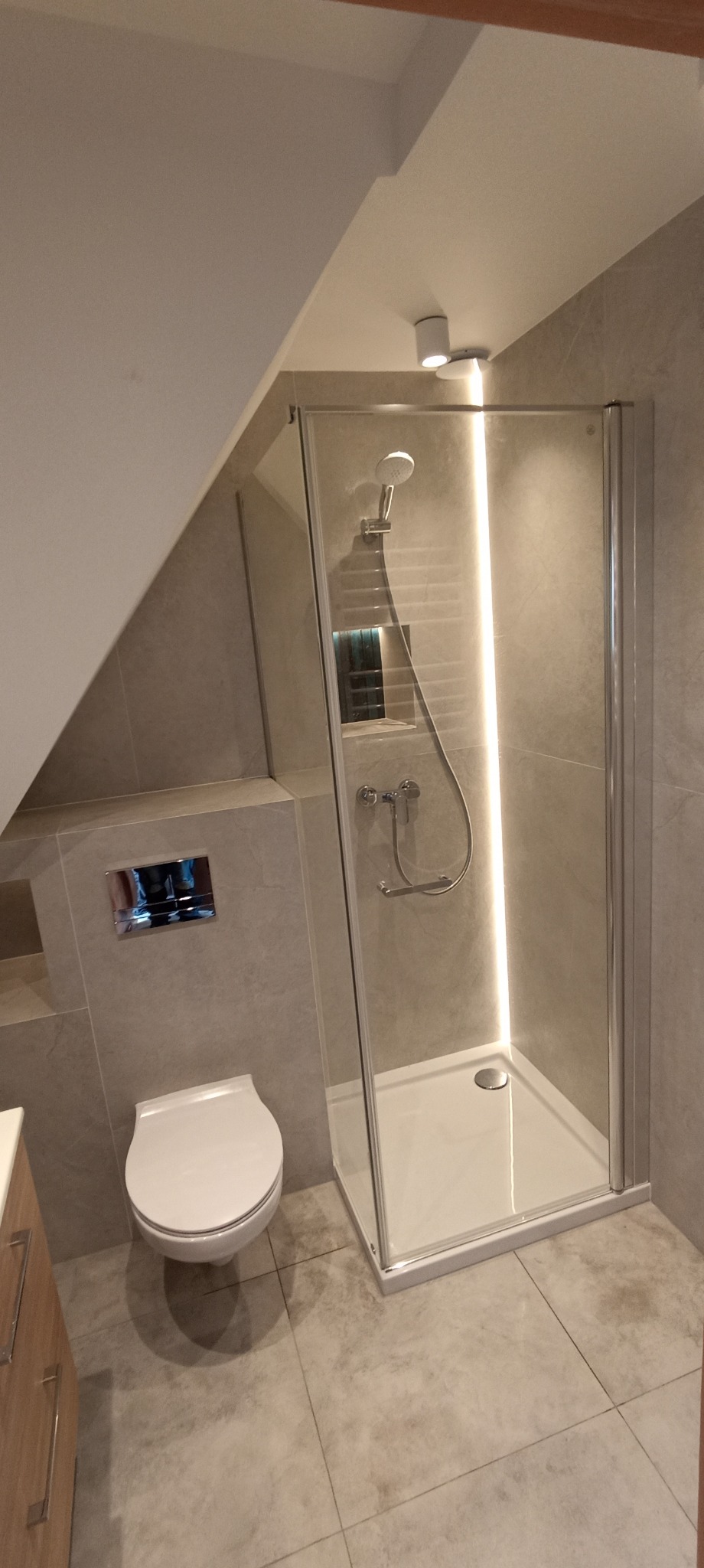 Wykończona łazienka na poddaszu ze szklaną kabiną prysznicową z podświetleniem LED i podwieszaną toaletą. Szare płytki podłogowe i ścienne.